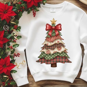 Könnte beinhalten: Weißes Sweatshirt mit einem Weihnachtsbaum-Design. Der Baum besteht aus geschichteten Abschnitten in roten, grünen und cremefarbenen Mustern, gekrönt von einer roten Schleife und einem goldenen Stern. Das Design hat eine rustikale Patchwork-Ästhetik.
