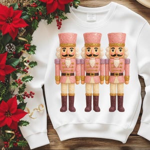 Könnte beinhalten: Weißes Sweatshirt mit drei rosa Nussknacker-Figuren. Die Nussknacker haben goldene Akzente, schwarze Schnurrbärte und braune Stiefel. Ein Weihnachtskranz mit roten Weihnachtssternen schmückt den Hintergrund.