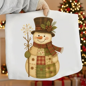 Puede incluir: Un artículo de tela blanca con un diseño de muñeco de nieve. El muñeco de nieve lleva un sombrero de copa marrón, una bufanda a cuadros y sostiene una varita de copo de nieve. El cuerpo del muñeco de nieve está hecho de tela de retazos.