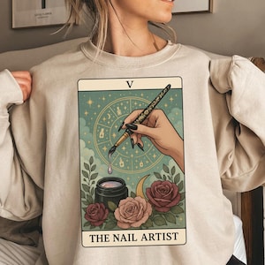May include: Beige sweatshirt featuring a tarot card design. The card depicts a hand holding a nail art brush, with a jar of polish, roses, and the text "THE NAIL ARTIST". The background includes a celestial design with stars and a crescent moon.