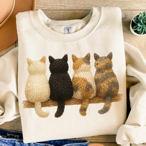 Könnte beinhalten: Cremefarbenes Sweatshirt mit einer Illustration von vier Katzen, die auf einem Ast sitzen. Die Katzen sind von hinten zu sehen, mit Farben wie Creme, Schwarz, Schildpatt und Braun-Grau. Das Design ist mittig auf der Vorderseite platziert.