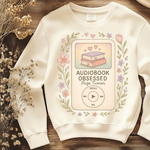 May include: Cream-colored sweatshirt with a floral design and the text "Audiobook Obsessed Page Turner." The graphic features a stack of books and a music player interface. The design includes pink, purple, and green accents.
