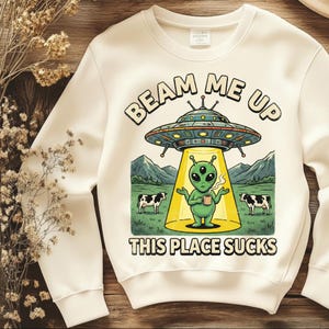 May include: Cream-colored sweatshirt with a graphic featuring a green alien holding a mug under a UFO. The text "BEAM ME UP" and "THIS PLACE SUCKS" are printed above and below the alien illustration. Two cows are also in the image.