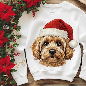 Peut inclure: Sweat-shirt blanc avec une illustration détaillée d'un chien brun et beige portant un bonnet de Père Noël rouge avec une bordure et un pompon blancs. Le visage du chien est centré sur le t-shirt. Article de vacances festif.