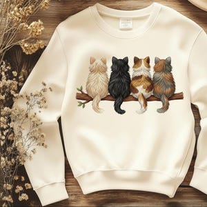 Könnte beinhalten: Cremefarbenes Sweatshirt mit einer Grafik von vier Katzen, die auf einem Ast sitzen. Die Katzen sind von hinten zu sehen, mit Fell in verschiedenen Farben: Beige, Schwarz, Braun und Weiß sowie Grau. Der Ast ist braun mit grünen Blättern.