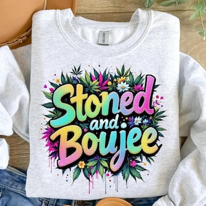 May include: A light gray sweatshirt with the words "Stoned and Boujee" in a colorful, gradient font. The text is surrounded by a wreath of cannabis leaves, flowers, and colorful accents. The sweatshirt is made of a soft material.