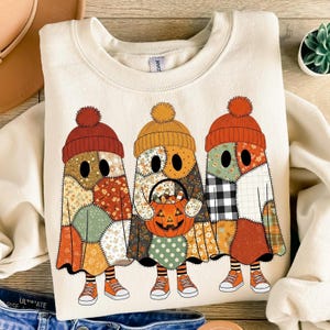 Puede incluir: Sudadera color crema con tres personajes fantasma con diseños de patchwork. Cada fantasma lleva un gorro de punto con pompón y zapatillas naranjas. Un fantasma sostiene un cubo de caramelos con forma de calabaza, perfecto para Halloween.