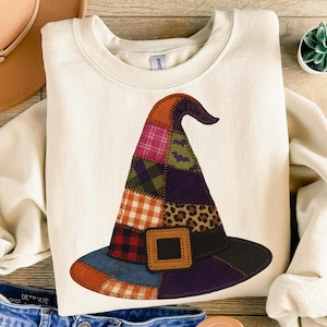May include: Cream-colored sweatshirt featuring a colorful patchwork witch hat design. The hat includes various patterns like plaid, leopard print, and bat silhouettes, with a brown buckle. A festive and cozy apparel item.