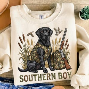 May include: Cream-colored sweatshirt featuring a black Labrador wearing a camouflage vest, sitting in front of tall grass and cattails. A rifle and backpack are next to the dog. The text "SOUTHERN BOY" is printed at the bottom.