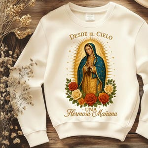 May include: Cream-colored sweatshirt featuring a religious illustration of a woman with a blue veil and gold halo, surrounded by roses. The text "Desde el Cielo" and "Una Hermosa Mañana" are printed in gold.