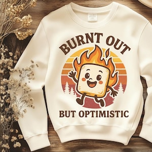 May include: Cream-colored sweatshirt with the words "BURNT OUT" above a cartoon marshmallow on fire, and the words "BUT OPTIMISTIC" below. The marshmallow has a smiling face and is set against a sunset-colored background.