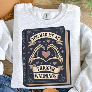 May include: Light gray sweatshirt featuring a graphic of a dark blue book with the text "YOU HAD ME AT TRIGGER WARNINGS." The book illustration includes skeleton hands forming a heart shape, surrounded by hearts.