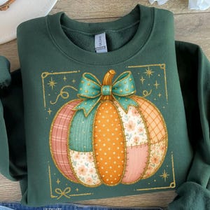 May include: A dark green sweatshirt featuring a colorful, patchwork pumpkin design. The pumpkin has a teal bow and is surrounded by a gold glitter frame with stars and a bow at the bottom. The pumpkin is made of various patterned fabrics.
