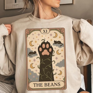 May include: Beige sweatshirt featuring a tarot card design with a cat paw, moons, stars, and the text "THE BEANS." The card has a decorative border and the Roman numeral "XI" at the top. The design is in shades of brown, beige, and green.