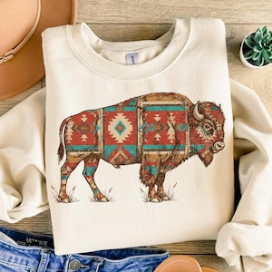 May include: Cream-colored sweatshirt featuring a detailed illustration of a bison. The bison's body is adorned with a Southwestern-inspired pattern in shades of red, turquoise, and brown. The sweatshirt has a crew neck and long sleeves, perfect for casual wear.