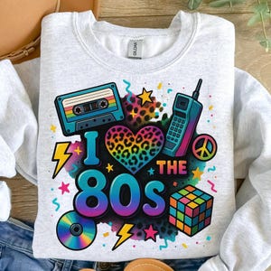 May include: A light grey sweatshirt featuring a vibrant 80s-themed design. The graphic includes a cassette tape, a mobile phone, a Rubik's cube, and the text "I ❤️ THE 80S" with a rainbow leopard print heart.