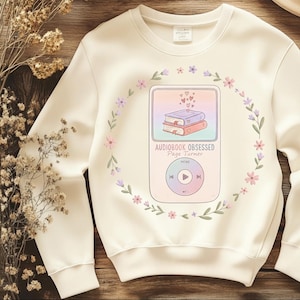 May include: Cream-colored sweatshirt with a floral wreath design. The graphic features a pastel-colored MP3 player with two books and the text "AUDIOBOOK OBSESSED Page Turner". The wreath includes pink and purple flowers.
