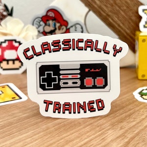 Könnte beinhalten: Ein weißer Aufkleber mit einem pixeligen Videospiel-Controller und dem roten Text "CLASSICALLY TRAINED". Weitere Aufkleber sind ein roter Pilz, ein gelbes Fragezeichen, ein gelber Stern und eine Mario-Figur.