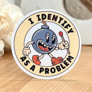 Puede incluir: Una pegatina redonda con un personaje de bomba de dibujos animados que sostiene una baqueta. El texto en la pegatina dice "Me identifico como un problema".