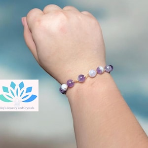 May include: A beaded bracelet featuring amethyst and moonstone beads strung on gold-colored wire. The bracelet is worn on a wrist. The image also includes a logo with the text "Joy's Jewelry and Crystals".