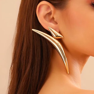 May include: Gold-colored statement earrings. The earrings feature a triangular stud and a long, curved, pointed design that extends down the neck. The earrings have a shiny, metallic finish and are worn on the ear.