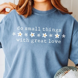 Puede incluir: Una camiseta azul jean con el texto "do small things with great love" y una fila de flores blancas con centros amarillos.
