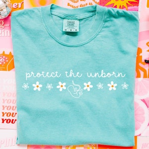 Puede incluir: Una camiseta azul claro con el texto "protect the unborn" en letras blancas y una fila de flores blancas. La camiseta está sobre un fondo colorido con otros artículos.