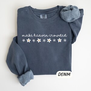K&ouml;nnte beinhalten: Ein denimblaues Sweatshirt mit dem wei&szlig;en Schriftzug "make heaven crowded" und einer Reihe wei&szlig;er G&auml;nsebl&uuml;mchen mit gelben Zentren. Das Sweatshirt mit Rundhalsausschnitt hat lange &Auml;rmel, von denen einer hochgekrempelt ist. Das Wort "DENIM" steht in einem wei&szlig;en Rechteck.