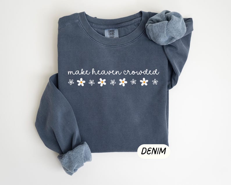 K&ouml;nnte beinhalten: Ein denimblaues Sweatshirt mit dem wei&szlig;en Schriftzug "make heaven crowded" und einer Reihe wei&szlig;er G&auml;nsebl&uuml;mchen mit gelben Zentren. Das Sweatshirt mit Rundhalsausschnitt hat lange &Auml;rmel, von denen einer hochgekrempelt ist. Das Wort "DENIM" steht in einem wei&szlig;en Rechteck.