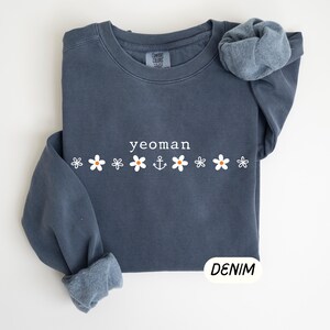 May include: A denim blue sweatshirt with the word "yeoman" in white, above a row of white flowers and an anchor graphic. The sweatshirt has a crew neck and long sleeves with rolled-up cuffs. The word "DENIM" is in a white oval.