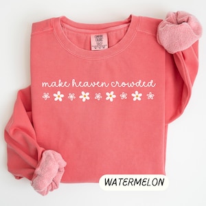 K&ouml;nnte beinhalten: Ein korallenfarbenes Sweatshirt mit dem Schriftzug "make heaven crowded" in wei&szlig;er Schrift, verziert mit wei&szlig;en G&auml;nsebl&uuml;mchen-Grafiken. Das Wort "WATERMELON" wird in einem wei&szlig;en Rechteck unten angezeigt. Die &Auml;rmel sind hochgekrempelt und zeigen ein rosa, flauschiges Innenfutter.