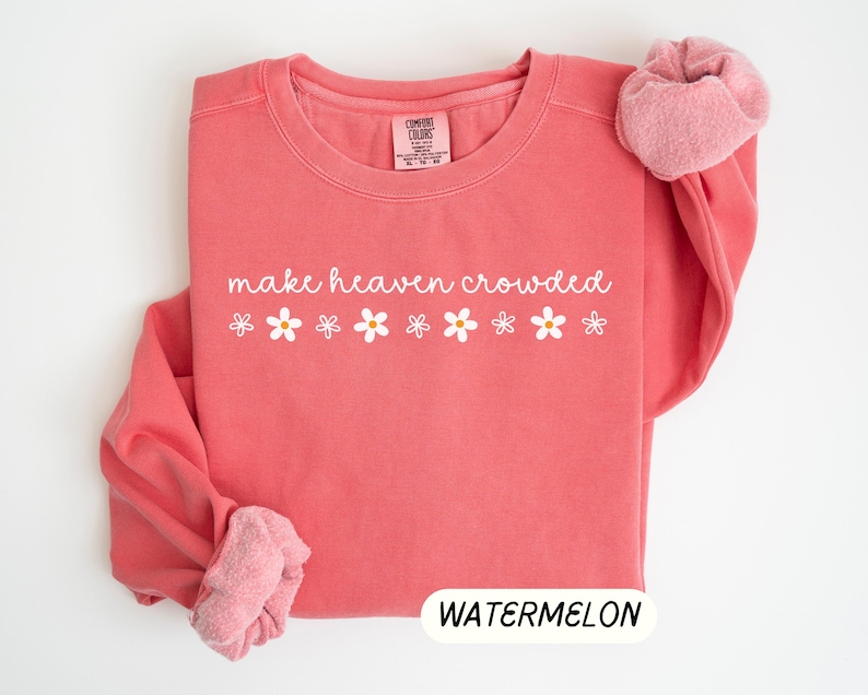 K&ouml;nnte beinhalten: Ein korallenfarbenes Sweatshirt mit dem Schriftzug "make heaven crowded" in wei&szlig;er Schrift, verziert mit wei&szlig;en G&auml;nsebl&uuml;mchen-Grafiken. Das Wort "WATERMELON" wird in einem wei&szlig;en Rechteck unten angezeigt. Die &Auml;rmel sind hochgekrempelt und zeigen ein rosa, flauschiges Innenfutter.