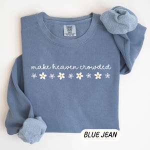 K&ouml;nnte beinhalten: Blaues Jeans-Sweatshirt mit dem Schriftzug "make heaven crowded" in wei&szlig;er Schrift und wei&szlig;en G&auml;nsebl&uuml;mchen-Grafiken. Das Sweatshirt hat einen Rundhalsausschnitt und lange &Auml;rmel. Die Worte "BLUE JEAN" stehen in einem wei&szlig;en Rechteck unten.