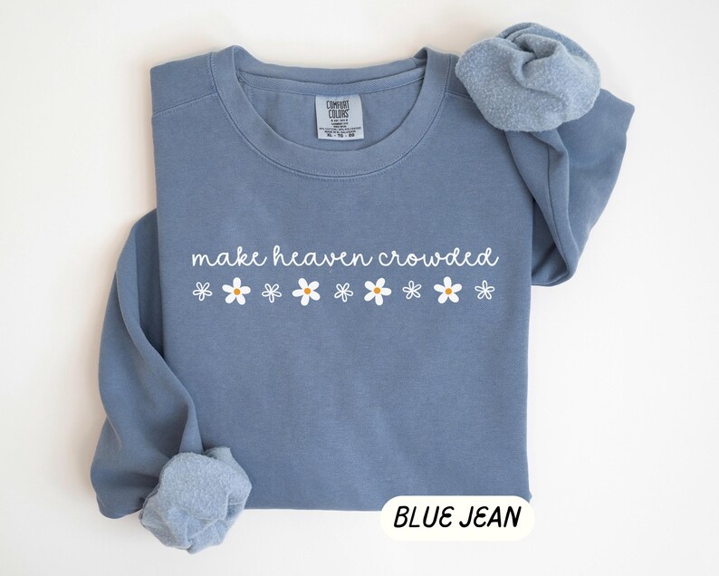 K&ouml;nnte beinhalten: Blaues Jeans-Sweatshirt mit dem Schriftzug "make heaven crowded" in wei&szlig;er Schrift und wei&szlig;en G&auml;nsebl&uuml;mchen-Grafiken. Das Sweatshirt hat einen Rundhalsausschnitt und lange &Auml;rmel. Die Worte "BLUE JEAN" stehen in einem wei&szlig;en Rechteck unten.