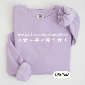 K&ouml;nnte beinhalten: Ein helllila Sweatshirt mit dem wei&szlig;en Schriftzug "make heaven crowded" und sechs wei&szlig;en G&auml;nsebl&uuml;mchen. Das Wort "ORCHID" ist in einem wei&szlig;en Rechteck unten aufgedruckt. Das Sweatshirt ist aus weichem Material.
