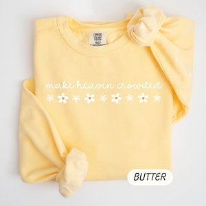 K&ouml;nnte beinhalten: Hellgelbes Sweatshirt mit dem wei&szlig;en Schriftzug "make heaven crowded" und sechs wei&szlig;en G&auml;nsebl&uuml;mchen-Grafiken. Das Wort "BUTTER" befindet sich in einem wei&szlig;en Oval unten.