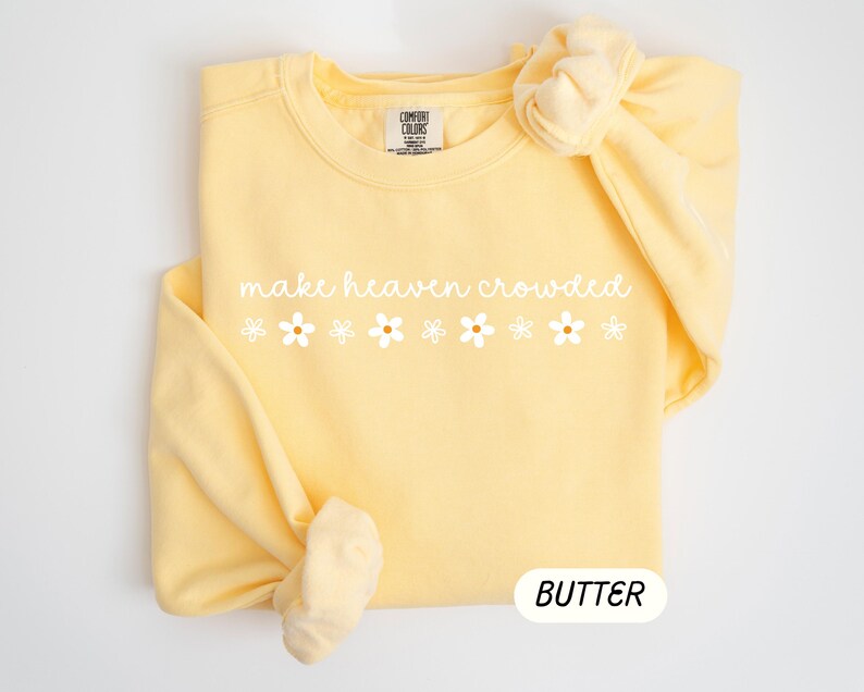 K&ouml;nnte beinhalten: Hellgelbes Sweatshirt mit dem wei&szlig;en Schriftzug "make heaven crowded" und sechs wei&szlig;en G&auml;nsebl&uuml;mchen-Grafiken. Das Wort "BUTTER" befindet sich in einem wei&szlig;en Oval unten.