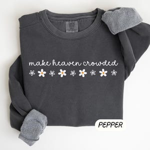 K&ouml;nnte beinhalten: Dunkelgraues Sweatshirt mit dem wei&szlig;en Schriftzug "make heaven crowded" und wei&szlig;en G&auml;nsebl&uuml;mchen-Grafiken. Die &Auml;rmel sind mit einem grauen, flauschigen Material versehen. Das Wort "PEPPER" ist wei&szlig; auf einem schwarzen Rechteck gedruckt.