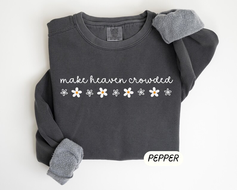 K&ouml;nnte beinhalten: Dunkelgraues Sweatshirt mit dem wei&szlig;en Schriftzug "make heaven crowded" und wei&szlig;en G&auml;nsebl&uuml;mchen-Grafiken. Die &Auml;rmel sind mit einem grauen, flauschigen Material versehen. Das Wort "PEPPER" ist wei&szlig; auf einem schwarzen Rechteck gedruckt.