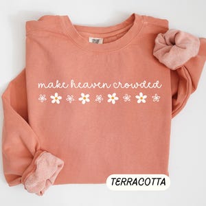 K&ouml;nnte beinhalten: Terracottafarbenes Sweatshirt mit dem wei&szlig;en Schriftzug "make heaven crowded" und wei&szlig;en G&auml;nsebl&uuml;mchen-Grafiken. Ein passendes Scrunchie liegt auf dem &Auml;rmel. Das Wort "TERRACOTTA" wird unten angezeigt.
