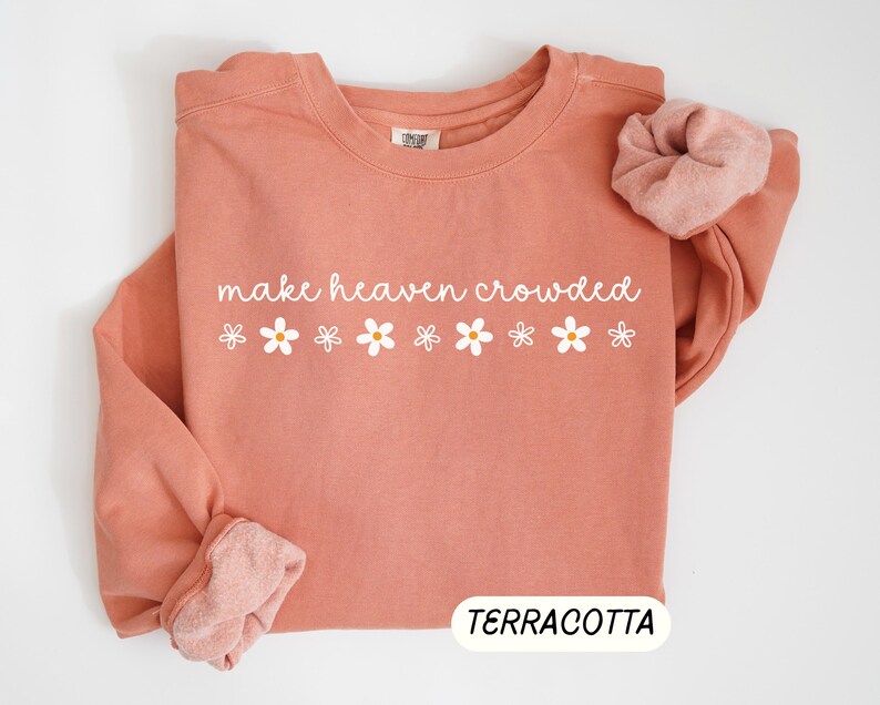 K&ouml;nnte beinhalten: Terracottafarbenes Sweatshirt mit dem wei&szlig;en Schriftzug "make heaven crowded" und wei&szlig;en G&auml;nsebl&uuml;mchen-Grafiken. Ein passendes Scrunchie liegt auf dem &Auml;rmel. Das Wort "TERRACOTTA" wird unten angezeigt.