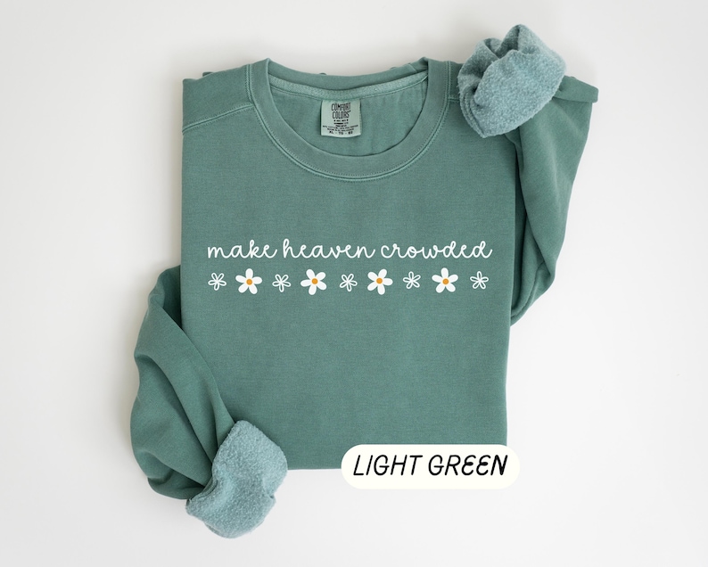 K&ouml;nnte beinhalten: Hellgr&uuml;nes Sweatshirt mit dem wei&szlig;en Schriftzug "make heaven crowded" und wei&szlig;en G&auml;nsebl&uuml;mchen. Die &Auml;rmel sind gefaltet. Unten rechts befindet sich ein Etikett mit der Aufschrift "LIGHT GREEN".