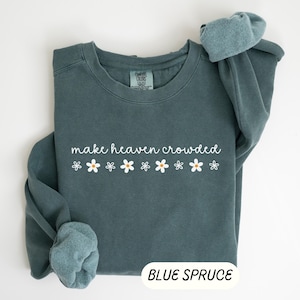 K&ouml;nnte beinhalten: Ein blau-fichtefarbener Sweatshirt mit dem wei&szlig;en Schriftzug "make heaven crowded", begleitet von wei&szlig;en G&auml;nsebl&uuml;mchen mit gelben Zentren. Das Sweatshirt hat einen Rundhalsausschnitt und lange &Auml;rmel.