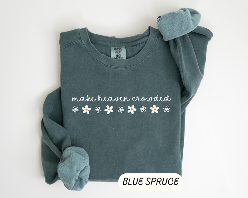K&ouml;nnte beinhalten: Ein blau-fichtefarbener Sweatshirt mit dem wei&szlig;en Schriftzug "make heaven crowded", begleitet von wei&szlig;en G&auml;nsebl&uuml;mchen mit gelben Zentren. Das Sweatshirt hat einen Rundhalsausschnitt und lange &Auml;rmel.