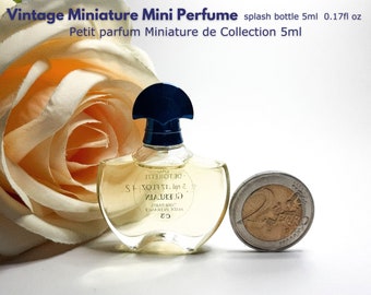 May include: A clear glass bottle of Guerlain perfume with a blue cap. The bottle is labeled "Guerlain" and "De Toilette" and contains 5 ml (0.17 fl oz) of perfume. The bottle is sitting on a white surface next to a 2 Euro coin.