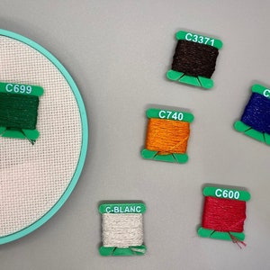 May include: A close-up of embroidery floss spools in various colors, including green, brown, orange, blue, red, and white. The spools are labeled with color codes, such as "C699", "C3371", "C740", "C820", "C600", and "C-BLANC".