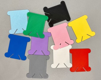 Könnte beinhalten: Sortiment von zehn Garnrollen in verschiedenen Farben, darunter Blau, Grün, Lila, Rosa, Gelb, Rot, Schwarz, Weiß und Grau. Die Spulen sind aus Kunststoff und haben eine rechteckige Form mit einem kleinen Loch in der Mitte.
