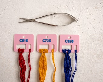 May include: Three pink plastic floss bobbins with red, yellow, and blue embroidery floss. The bobbins are labeled with the numbers C816, C725, and C820. A pair of silver embroidery scissors is above the bobbins.