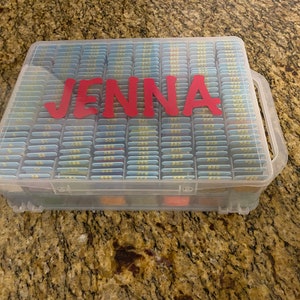 May include: A clear plastic storage box with a handle, filled with small, labeled containers. The box has the name "JENNA" written on the front in red letters.