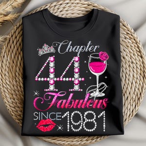 May include: Black t-shirt with a graphic design celebrating a 44th birthday. The design includes a tiara, the words "Chapter 44 Fabulous Since 1981", a wine glass with a rose, and a pair of red lips.